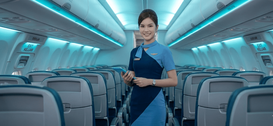 Foto detail pramugari Indonesia usia muda berdiri di terminal bandara modern. Fokus pada syal leher biru muda yang diikat rapi, lencana sayap emas kecil di dada kiri, dan kancing seragam yang elegan. Di tangannya terdapat pegangan koper hitam besar.